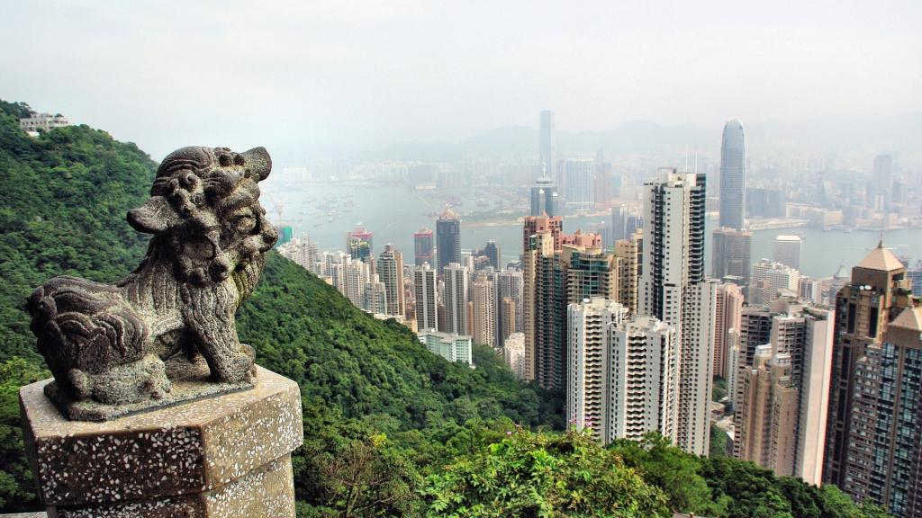 A traditional lion sculpture on a lush hillside overlooking a coastal city dense with high-rises.